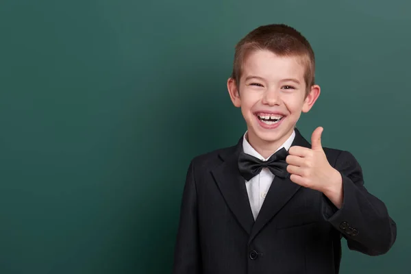 School boy show best gesture, portrait near green blank chalkboard background, dressed in classic black suit, one pupil, education concept — Stock Photo, Image