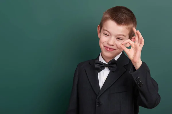 School jongen Toon ok teken, portret in de buurt van groene leeg schoolbord achtergrond, gekleed in een klassieke zwarte pak, één leerling, onderwijs concept — Stockfoto