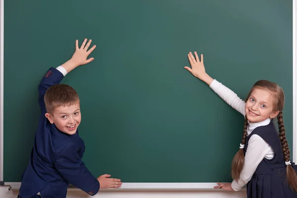 Niño y niña de la escuela primaria poner las manos en el fondo de pizarra y mostrar el espacio en blanco, vestido con traje negro clásico, alumno de grupo, concepto de educación — Foto de Stock