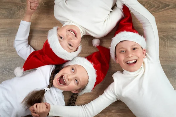 Tres niños en los sombreros de Santa que mienten en el fondo de madera, teniendo emociones divertidas y felices, concepto de vacaciones de invierno —  Fotos de Stock