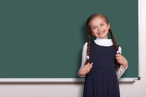 Szép iskolás lány a pigtail mosolygott közelében üres Palatábla háttér, klasszikus fekete színben, oktatási koncepció öltözött — Stock Fotó