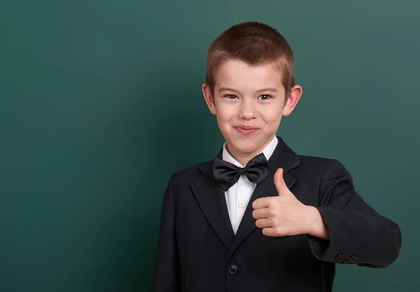 School boy show best gesture, portrait near green blank chalkboard background, dressed in classic black suit, one pupil, education concept — Stock Photo, Image