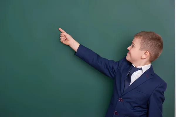 Chico de la escuela señalar el dedo cerca de fondo pizarra en blanco, vestido con traje negro clásico, alumno de grupo, concepto de educación — Foto de Stock