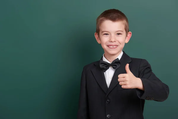 School boy show best gesture, portrait near green blank chalkboard background, dressed in classic black suit, one pupil, education concept — Stock Photo, Image