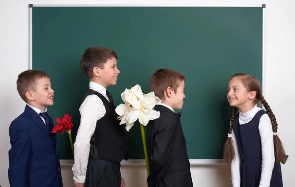 Ragazzi dando fiori ragazza, bambino della scuola elementare vicino sfondo lavagna vuota, vestito in classico abito nero, allievo di gruppo, concetto di istruzione — Foto Stock