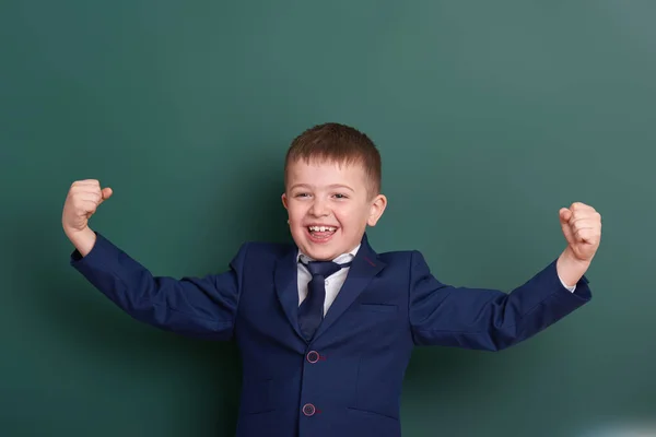 Niño de la escuela muestran músculos bíceps, retrato cerca de fondo de pizarra verde en blanco, vestido con traje negro clásico, un alumno, concepto de educación — Foto de Stock