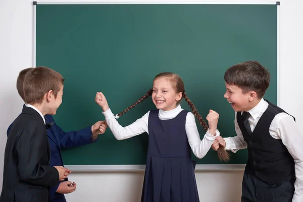 Dziecko gry i zabawy, chłopcy wyciągnąć dziewczynę warkocze, w pobliżu puste szkoła tablica tło, ubrany w klasyczny czarny garnitur, grupa uczniów, koncepcja edukacji — Zdjęcie stockowe