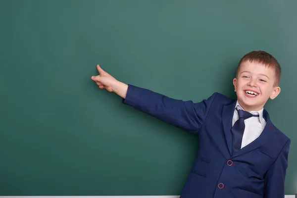 Chłopiec szkoły punkt palcem w pobliżu pusta tablica tło, ubrany w klasyczny czarny garnitur, grupa uczniów, koncepcja edukacji — Zdjęcie stockowe