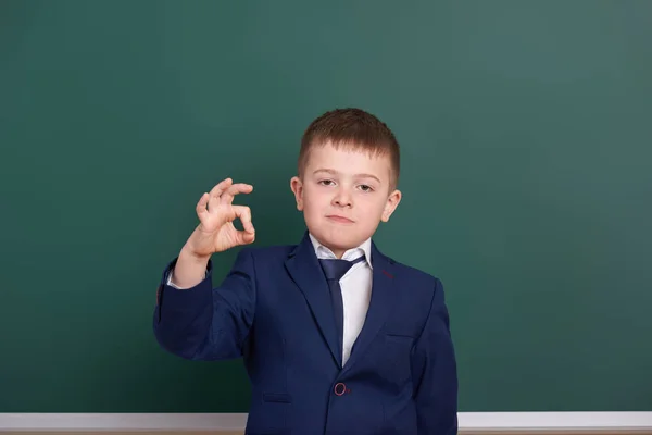 Škola chlapec Ukázat ok znamení, portrét poblíž pozadí zelené prázdnou tabuli, oblečený v klasickém černém obleku, jeden žák, koncepce vzdělávání — Stock fotografie