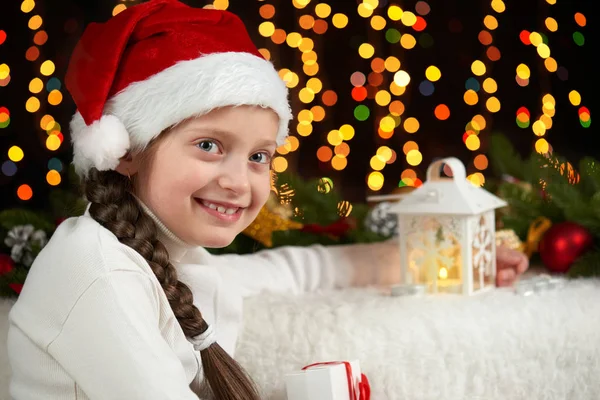 Bambino ragazza ritratto con decorazione natalizia, sfondo scuro con luci, espressione del viso ed emozioni felici, vestito in cappello di Babbo Natale, concetto di vacanza invernale — Foto Stock