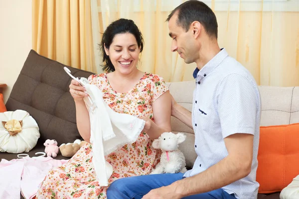 家长为刚出生的婴儿选择衣服。怀孕的女人和男人。幸福的夫妇坐在家里的沙发上 — 图库照片