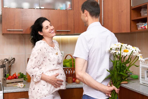 Muž dává heřmánek květiny na těhotnou ženu, pár v interiéru kuchyně s čerstvého ovoce a zeleniny, zdravé jídlo koncept — Stock fotografie