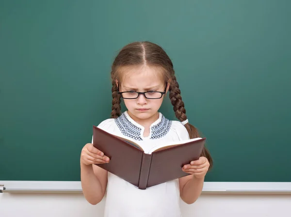 Schulmädchen lesen Buch, posieren an der Schultafel, leerer Raum, Bildungskonzept — Stockfoto