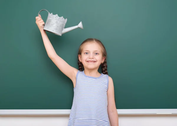 Schoolgirl with watering can play near a blackboard, empty space, education concept — Stock Photo, Image