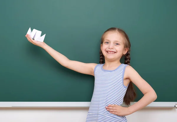 Schoolgirl with paper boat play near a blackboard, empty space, education concept — Stock Photo, Image