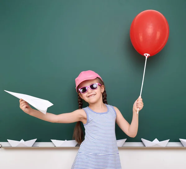 Schoolgirl with paper plane and balloon play near a blackboard, empty space, education concept — Stock Photo, Image