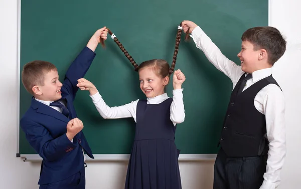 Child play and having fun, boys pull the girl braids, near blank school chalkboard background, dressed in classic black suit, group pupil, education concept — Stock Photo, Image