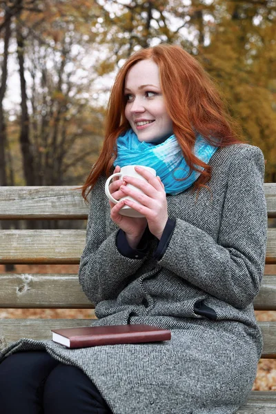 Redhead girl with cup in autumn park, yellow leaves and trees — Stock Photo, Image