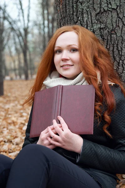 Giovane donna seduta vicino all'albero nel parco autunnale e leggere libro, foglie gialle — Foto Stock