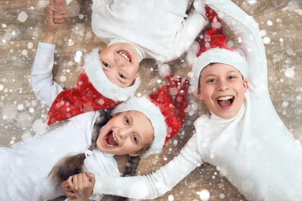 Счастливый ребенок, держась за руки и лежа на деревянном фоне, одетый в шапку Деда Мороза и веселящийся, концепция зимнего праздника, украшение снега — стоковое фото