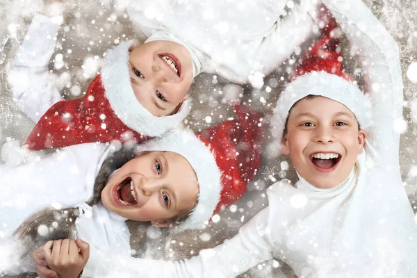 กลุ่มเด็กนอนด้วยกันบนพื้นหลังไม้และหัวเราะ แต่งตัวในหมวกคริสต์มาสซานต้าและมีความสนุกสนานแนวคิดวันหยุดฤดูหนาวตกแต่งหิมะ — ภาพถ่ายสต็อก
