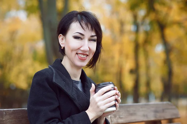 Jovem mulher sentada em um banco e beber café, estação de outono, parque da cidade, folhas amarelas — Fotografia de Stock