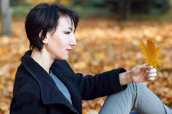 Girl in black coat sitting on a yellow leaves in autumn city park, fall season — Stock Photo, Image