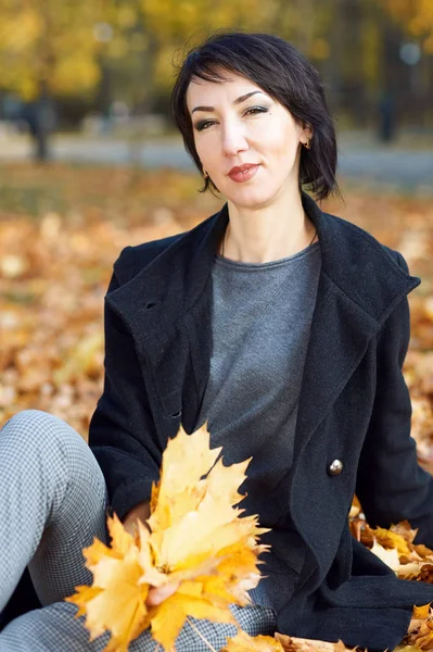 Girl in black coat sitting on a yellow leaves in autumn city park, fall season — Stock Photo, Image