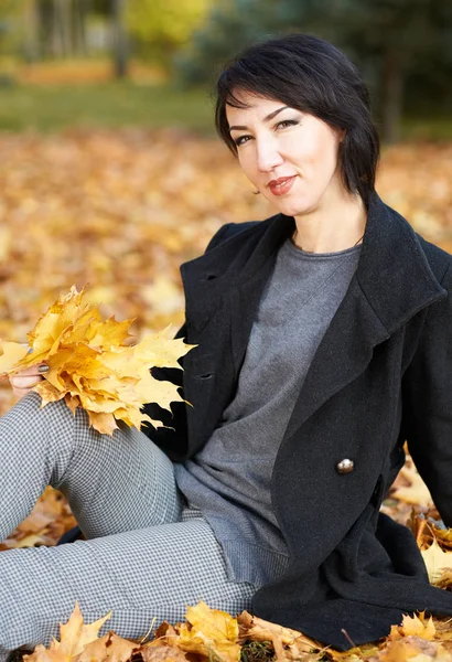 Girl in black coat sitting on a yellow leaves in autumn city park, fall season — Stock Photo, Image