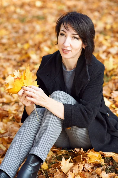 Girl in black coat sitting on a yellow leaves in autumn city park, fall season — Stock Photo, Image