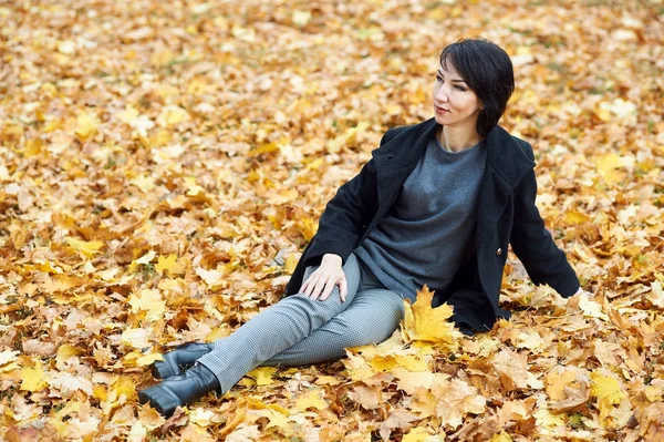 Dívka v černém kabátě, sedí na žluté listy v podzimní městského parku, podzim — Stock fotografie