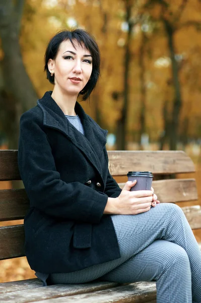 Junge Frau mit Kaffee auf einer Bank im herbstlichen Stadtpark, gelben Blättern und Bäumen — Stockfoto