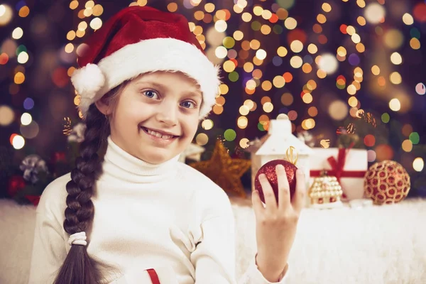 Menina criança posando com decoração de Natal no fundo escuro, luzes iluminadas e bokeh, close-up rosto, vestido com chapéu de santa, conceito de férias de inverno, árvore de xmas — Fotografia de Stock