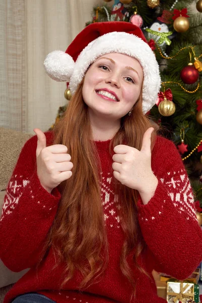 Dívka v retro červený svetr a čepice santa ukazují nejlepší gesto. Domácí interiér s vánoční výzdobou, jedle a dary. Nový rok eve a zimní dovolená concept. — Stock fotografie