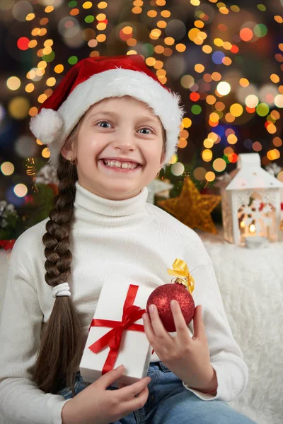 Menina criança vestida com chapéu de Papai Noel com presentes de Natal no fundo iluminado escuro, feliz ano novo e conceito de celebração de inverno — Fotografia de Stock