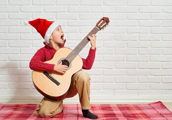 Chlapec hrající hudbu na kytaru, na sobě červený vlněný svetr a čepice santa, sedící na červené kostkované dece, bílé zdi na pozadí — Stock fotografie