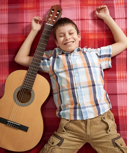 Menino tocando música na guitarra, encontra-se em um cobertor xadrez vermelho, vista superior — Fotografia de Stock