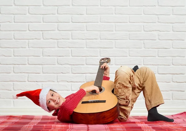 Menino tocando música na guitarra, vestido com uma camisola de lã vermelha e chapéu de santa, sentado em um cobertor xadrez vermelho, parede de tijolo branco no fundo — Fotografia de Stock