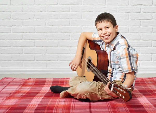 Boy playing music on guitar, sitting on a red checkered blanket, white brick wall background — Stock Photo, Image