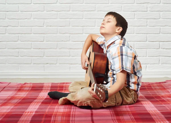 Chlapec hrající hudbu na kytaru, sedí na pozadí zdi cihlové červené kostkované dece, bílé — Stock fotografie