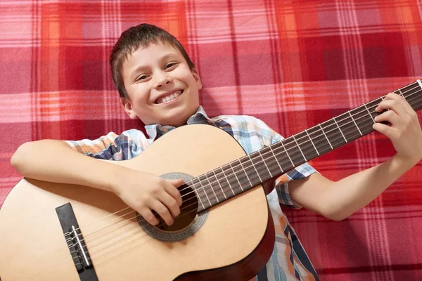 Boy playing music on guitar, lies on a red checkered blanket, top view — Stock Photo, Image