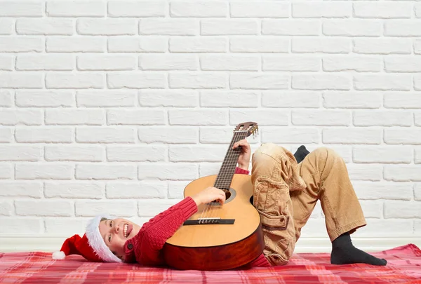 Chlapec hrající hudbu na kytaru, na sobě červený vlněný svetr a čepice santa, sedící na červené kostkované dece, bílé zdi na pozadí — Stock fotografie