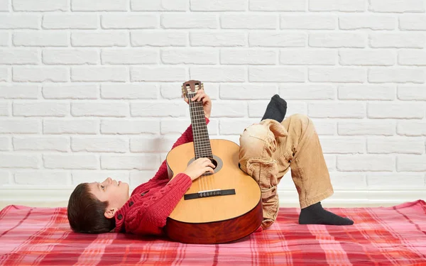 Chlapec hrající hudbu na kytaru, oblečený v červené vlněné svetry, leží na červené kostkované dece, bílé zdi na pozadí — Stock fotografie