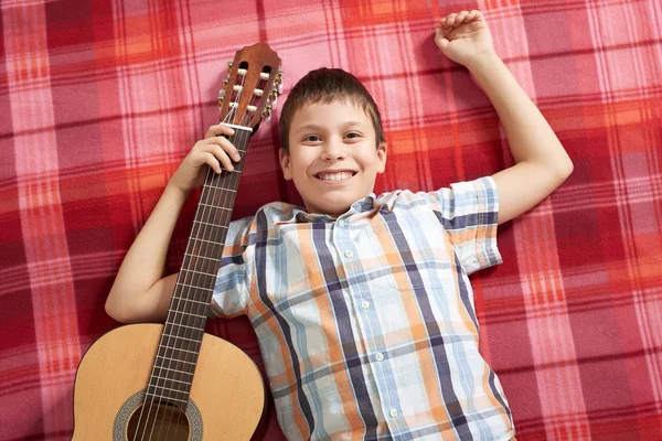 Menino tocando música na guitarra, encontra-se em um cobertor xadrez vermelho, vista superior — Fotografia de Stock