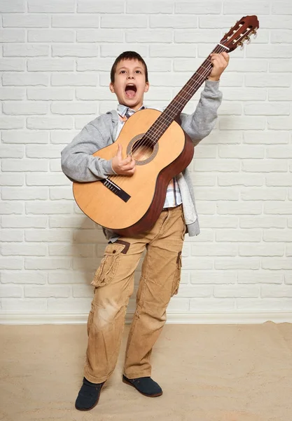 Chlapce hrát na kytaru, stojící v plné výšce, bílá cihlová zeď pozadí — Stock fotografie