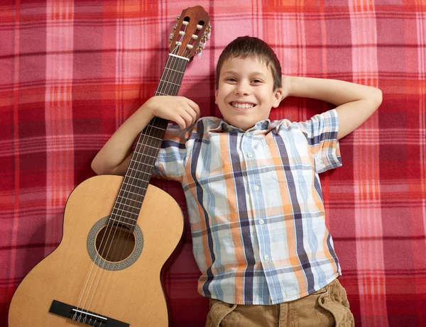 Menino tocando música na guitarra, encontra-se em um cobertor xadrez vermelho, vista superior — Fotografia de Stock