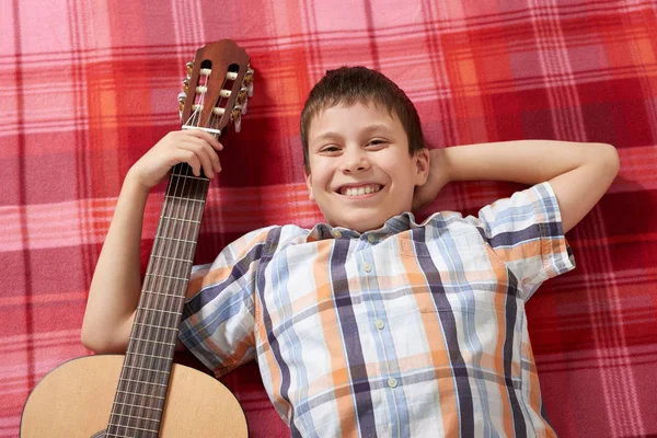 Boy playing music on guitar, lies on a red checkered blanket, top view — Stock Photo, Image