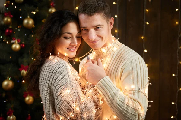 Giovane coppia insieme in luci di Natale e decorazione, vestito di bianco, abete su sfondo di legno scuro, serata romantica, concetto di vacanza invernale — Foto Stock