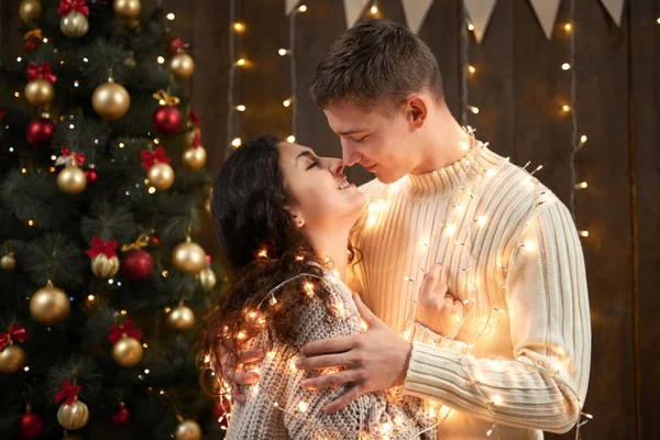 Jong koppel zoenen in Kerstverlichting en decoratie, gekleed in het wit, fir tree op donkere houten achtergrond, een romantische avond, een concept van winter vakantie — Stockfoto