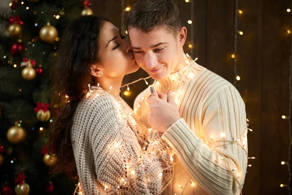 Jovem casal beijando em luzes de Natal e decoração, vestido de branco, abeto no fundo de madeira escura, noite romântica, conceito de férias de inverno — Fotografia de Stock